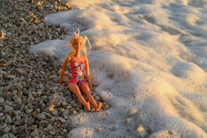 Lutka sjedi na pješčanoj plaži. Morski valovi zapljuskuju Barbie lutku.