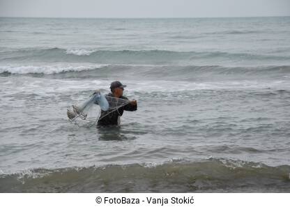 Muškarac stoji u uzburkalom moru i baca mrežu u vodu. Ribar pokušava da ulovi ribu tokom kišnog dana na moru.