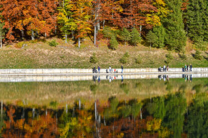 Ljudi šetaju pored vode tokom lijepog jesenjeg dana. Odsjaj crvenih, žutih i zelenih krošnji na površini vode.