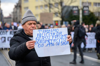 Stariji muškarac stoji na ulici tokom javnog protesta u Sarajevu i drži ručno ispisan transparent. Na papiru je poruka napisana plavim markerom. U pozadini se nalaze drugi učesnici protesta.