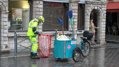 Čistač ulice. Muški čistač ulica sa kolicima za čišćenje. Čovjek u radnom odijelu prazni kante za smeće. Čišćenje ulice.
