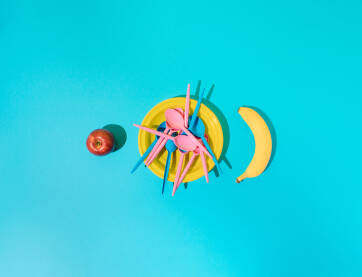 Žuti plastični tanjur s plavim i ružičastim plastičnim kašikama, bananom i jabukom.