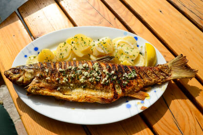 Riba grilovana na žaru u restoranu. Ukusna i slasna riba s krumpirom, limunom i češnjakom na tanjiru na drvenom stolu. Hrana.