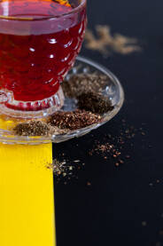 Staklena šoljica crvenog čaja na crno - žutoj postavi posutoj sa suhim travama čaja
