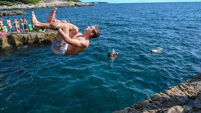 Mladić skače u more. Čovjek radi salto iznad vode. Mladi se zabavljaju na plaži i u moru.