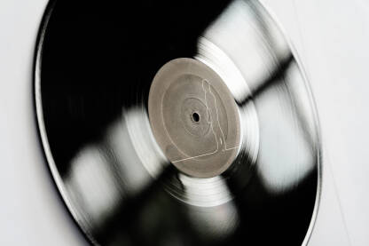 Ova slika prikazuje crno-belu vinilnu gramofonsku ploču na beloj pozadini. Ploča djeluje izgrebano i istrošeno, s brazdama koje su vidljive, na beloj pozadinom koja je u kontrastu s crno-belom pločom.