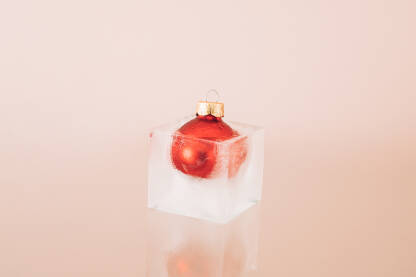 Crvena božićna kuglica / ukras zaleđena u kocki leda. Božićna / Novogodišnja čestitka.