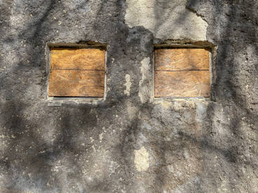 Zid stare, ruševne kuće sa dva prozora prekrivena daskama. Stara kuća u sjeni drveta.