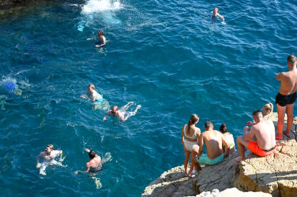 Mladi ljudi se ljeti zabavljaju na stjenovitoj plaži. Ljudi se sunčaju i kupaju u moru. Prepuna plaža. Ljetna sezona.