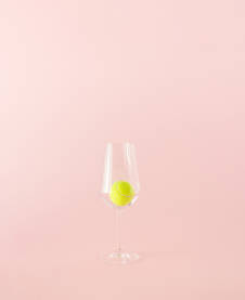 Žuta teniska loptica u vinskoj čaši na roze pozadini.