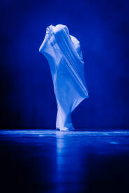 Žena u beloj haljini stoji na tamnoj sceni, dok joj haljina teče i leprša oko nje. Scena je osvetljena plavim svetlom. Njeno lice je skriveno haljinom dok joj telo ostaje vidljivo.