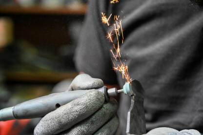 Radnik sa zaštitnim rukavicama koristi rotacijski alat za brušenje metala, pri čemu lete iskre. Industrijski radnik oblikuje metalni komad pomoću rotacijskog alata.