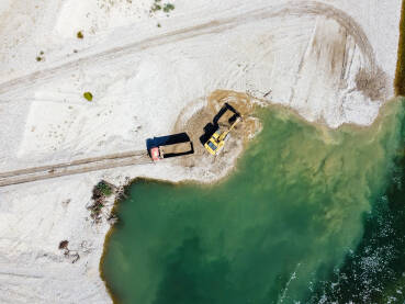Bager kopa i utovaruje pijesak u kiper na obali rijeke. Teška mehanizacija radi u kamenolomu pijeska. Snimak dronom na bager i kamion koji utovaruju šljunak u prirodi. Životna sredina i zagađenje vode