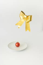 Crvena ukrasna kuglica na bijelom tanjuru i zlatna mašna u zraku.