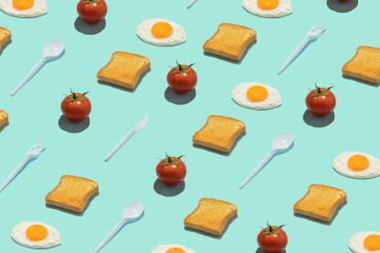 Kreativna pozadina s tostiranim kruhom, rajčicama, pečenim jajima i plastičnim priborom za jelo na plavoj pozadini.