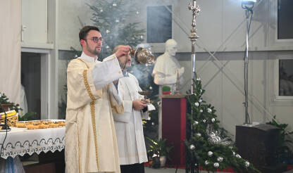 Katoličko svećenstvo u liturgijskom ruhu služi crkvenu misu u blizini božićnog drvca i kipa. Svećenici i đakoni slave božićnu misu.