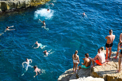 Mladi ljudi se ljeti zabavljaju na stjenovitoj plaži. Ljudi se sunčaju i kupaju u moru. Prepuna plaža. Ljetna sezona.