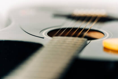 Fotografija prikazuje detaljan pogled na akustičnu gitaru. Gitara ima crno telo sa belom zaštitnom pločicom i belim štelujućim čivijama. Pozadina je bela površina sa svjetlosnim refleksijama.