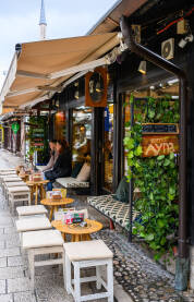 Kafić na Baščaršiji u Sarajevu, sa otvorenom baštomt, rustikalnim stolicama i prolaznicima koji uživaju u kafi. Autentična atmosfera starog grada.