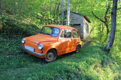 Stari narandžasti "fićo" (Zastava 750) parkiran uz šumsku stazu i okružen svježim proljetnim zelenilom. U pozadini se nazire mala kamena kovačnica.
