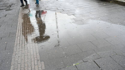 Refleksija osobe sa kišobranom u lokvi vode. Kiša na asfaltnom putu. Kapljice kiše u lokvi. Ljudi sa hodaju sa kišobranom.