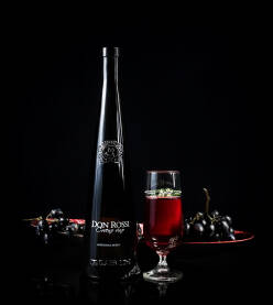 Elegantna flaša crnog vina, postavljene na stolu pored čaše sa crnim vinom i crnim grožđem u pozadini. Rubinovo crno/crveno vino Don Rossi.