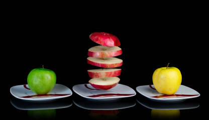 Plutajuća kriška crvene jabuke između zelene i žute jabuke na tanjirima.