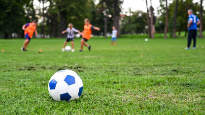 Djeca igraju fudbal u parku. Lopta u prvom planu dok se djeca igraju u pozadini na zelenoj travi. Sretna i slatka djeca koja igraju fudbalsku utakmicu na stadionu. Fudbalska utakmica.