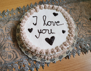 Okrugla torta ukrašena bijelom kremom i čokoladnom glazurom, s ispisanom porukom „Volim te“ i motivima srca na vrhu. Prikladna za teme ljubavi, romantike i posebnih prilika.