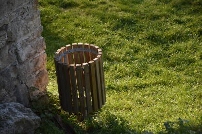 Drvena korpa za smeće pored zelene površine i kamene građevine.