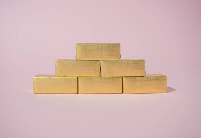 Čokolade umotane u papir zlatne boje naslagane u obliku piramide kao zlatne poluge na pastelnoj podlozi roze boje.