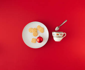 Keksi i božićna, novogodišnja kuglica na bijelom tanjuru i šalica za čaj s kašikicom na crvenoj pozadini.