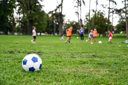 Djeca igraju fudbal na školskom terenu. Lopta u prvom planu dok se dječaci igraju u pozadini na zelenoj travi.