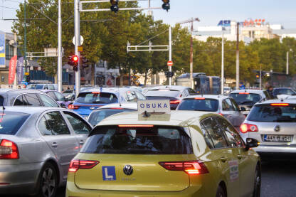 Žuti automobil s oznakom "OBUKA" stoji u koloni na semaforu, okružen drugim vozilima. Gužva u saobraćaju. Saobraćajni kolaps u gradu.