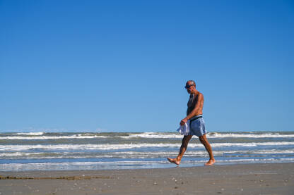 Čovjek šeta pješčanom plažom u blizini mora. Stariji muškarac uživa u šetnji uz more.