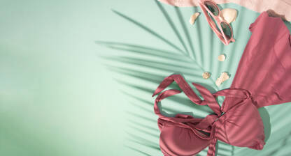 Ženski kupaći kostim, naočale, peškir pastelne pink boje i nekoliko bijelih školjkica sa sjeno palminog lista na svijetloj, tirkiznoj podlozi. Ljeto, plaža, bazen, sunce, kupanje, plivanje - koncept