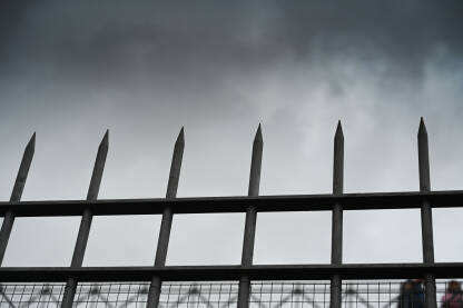 Šiljci ograde uzdižu se prema olujnim oblacima. Vrh metalne ograde naspram tamnog dramatičnog neba. Oštra metalna ograda pod teškim tamnim oblacima.